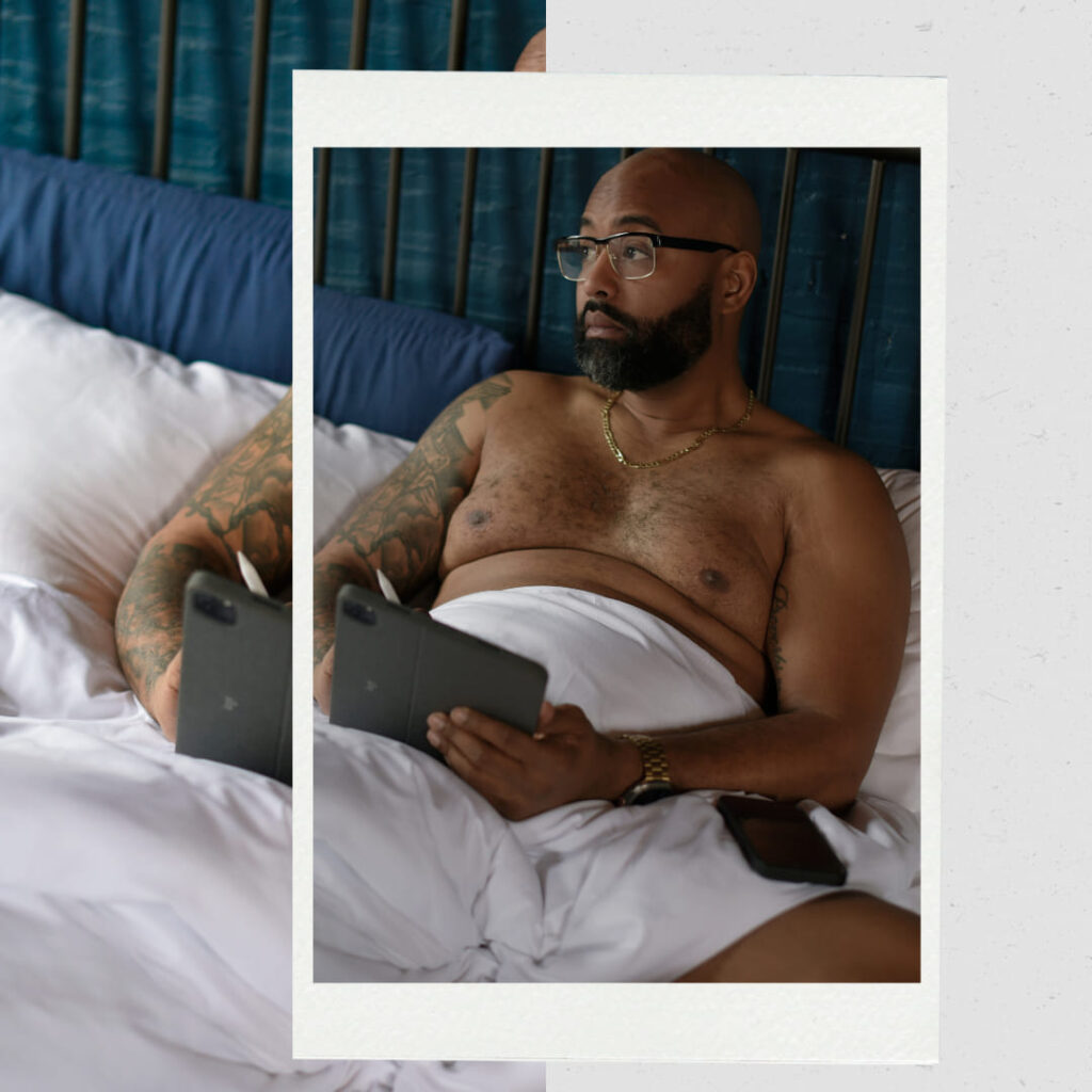 Black man laying in bed with white duvet cover with an iPad