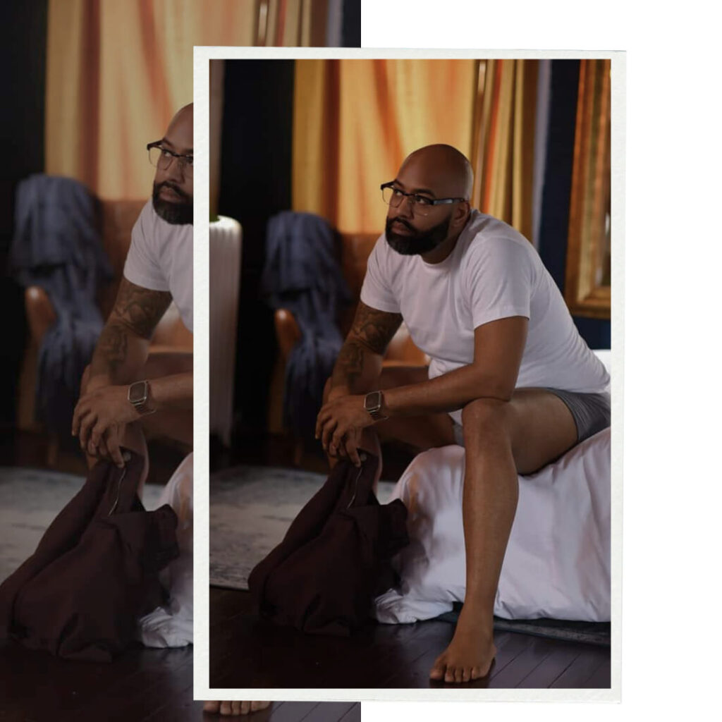 IMAGE of BLACK man in a white t shirt with grey boxer briefs, He is sitting down on bed with plum colored pants in his hand